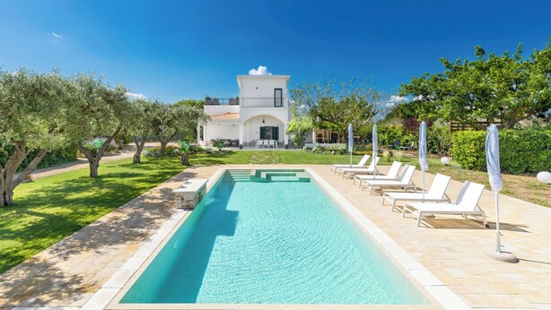 Una piscina con sillones de descanso, un césped cuidado y una casa al fondo.