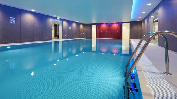 Una piscina interior con agua cristalina, una escalera de metal y una pared roja al fondo.