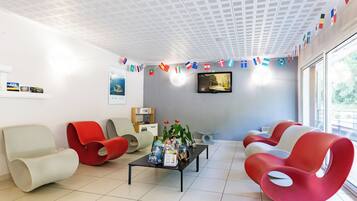Un espace d’attente moderne avec des chaises rouges et blanches, une table basse avec des magazines et un téléviseur fixé au mur.