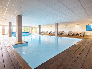 An indoor swimming pool with wooden decking, lounge chairs, and a tiled ceiling.