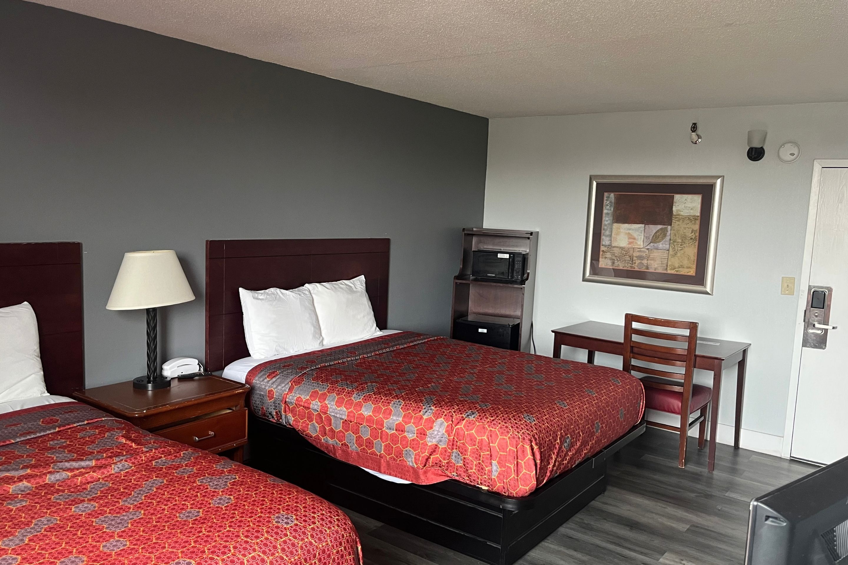 A hotel room with two beds, a desk, a chair, a lamp, a microwave, and a painting on the wall.