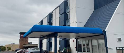 A modern building with a covered entrance, a parking lot with several cars, and a clear sky.