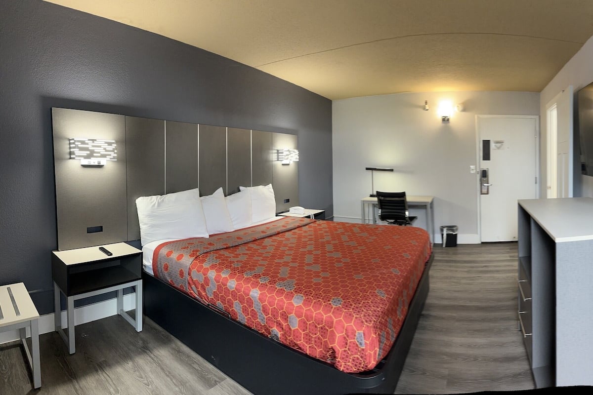 A modern hotel room with a large bed, a desk, a TV, and a small seating area.