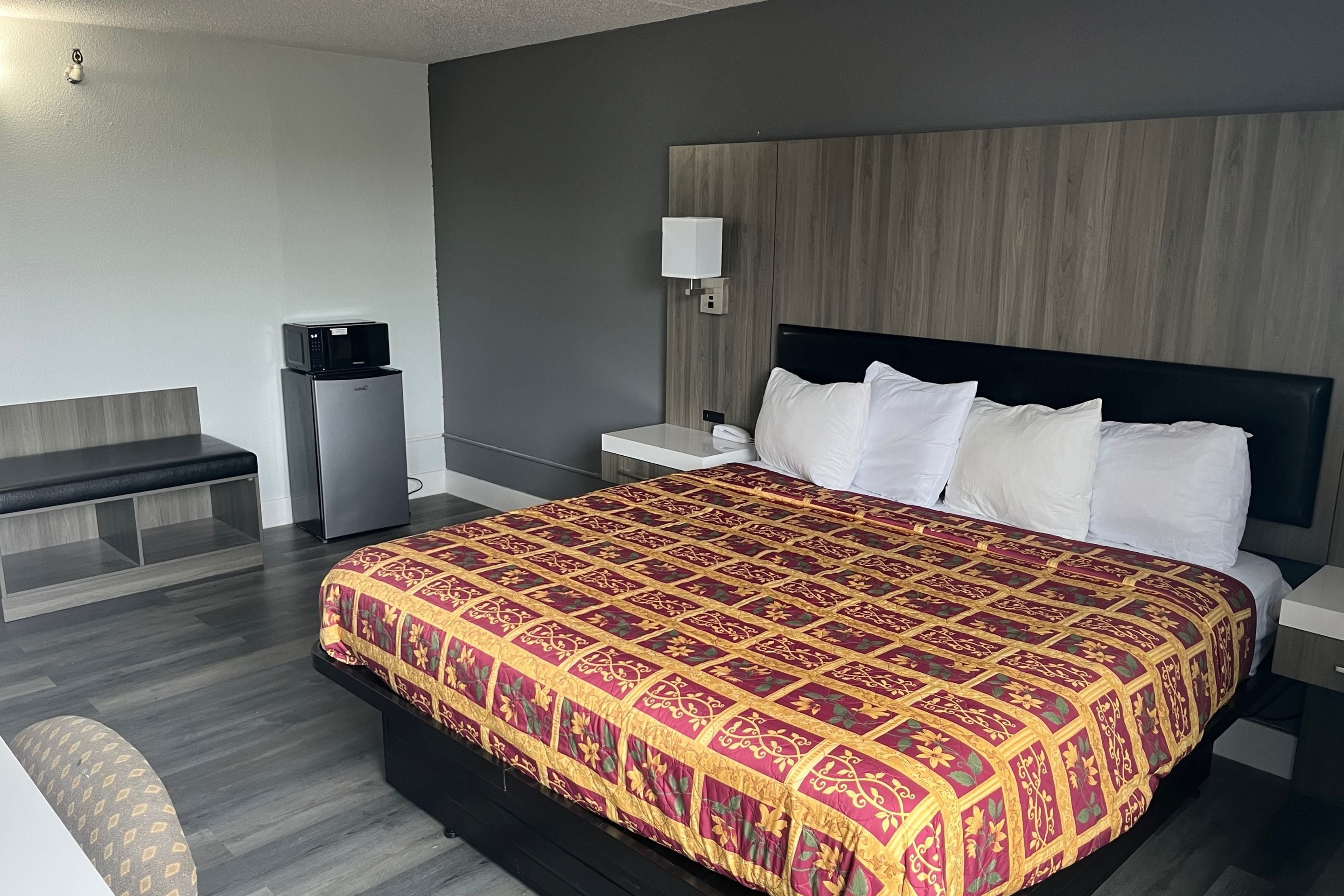 A hotel room with a large bed, a bedside table, a small refrigerator, a bench, and a wall-mounted lamp.