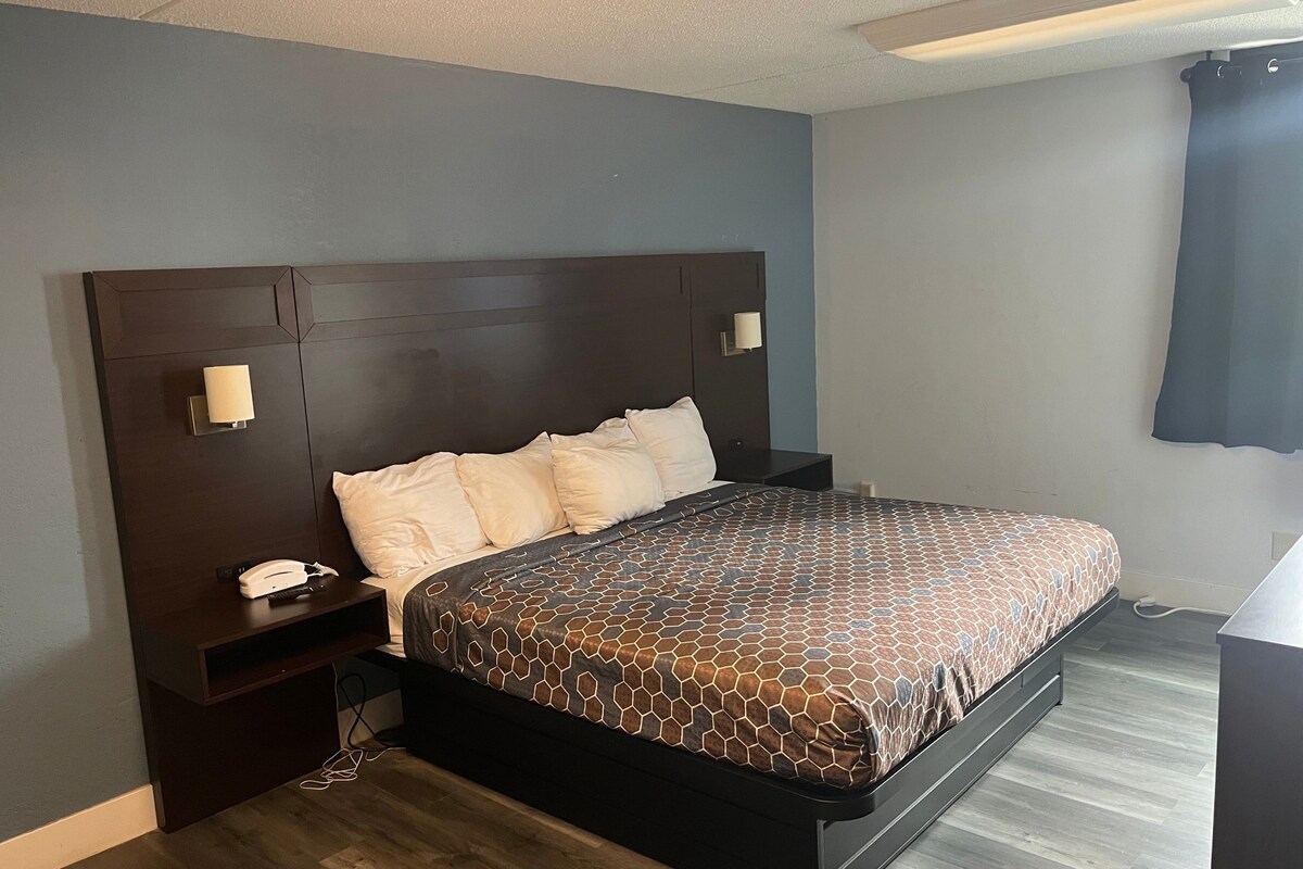 A hotel room with a dark wood headboard, a bed with a patterned bedspread, a bedside table with a phone and remote, and a wall-mounted light fixture.