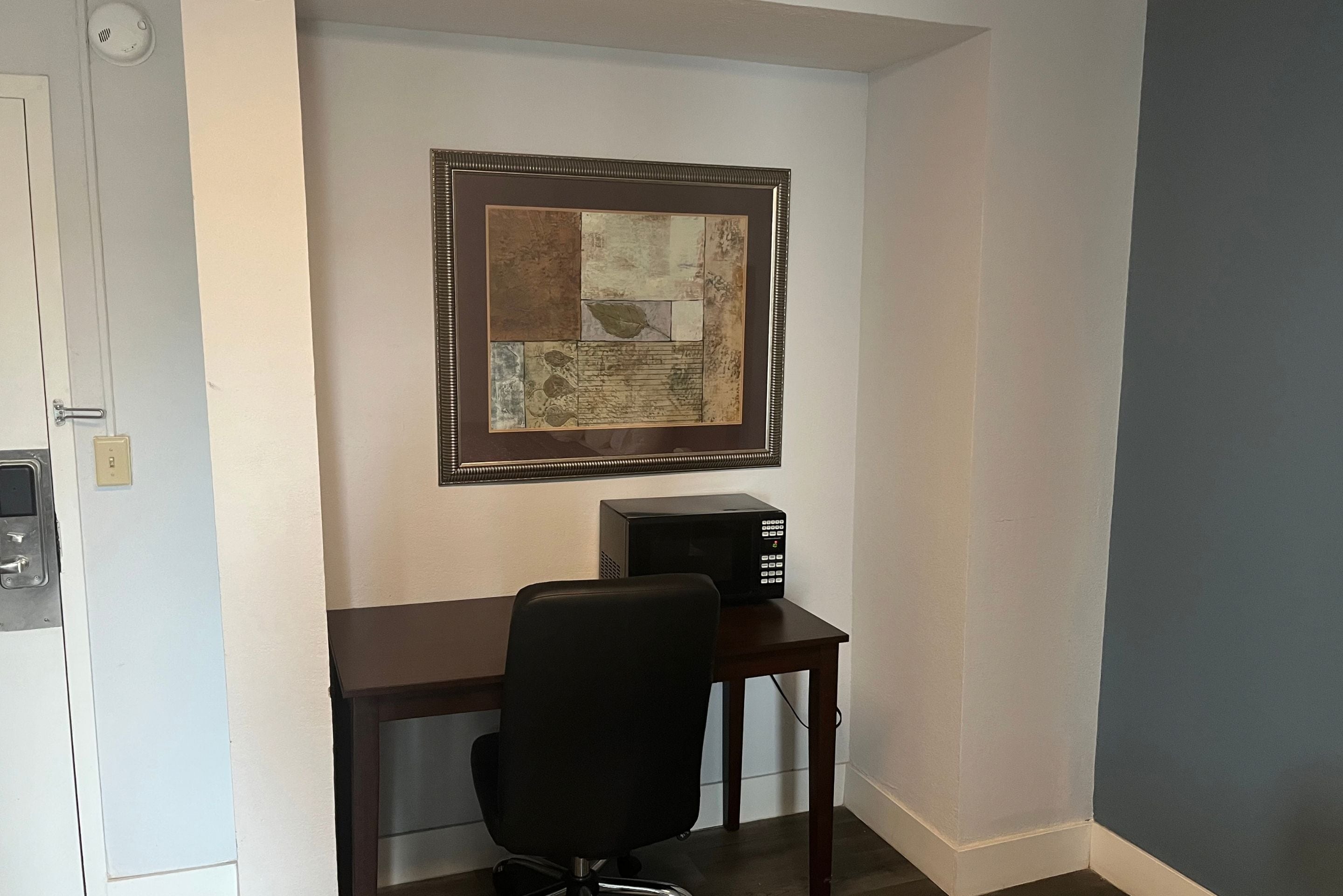 Un pequeño espacio de oficina con un escritorio, una silla, un microondas y una obra de arte enmarcada en la pared.