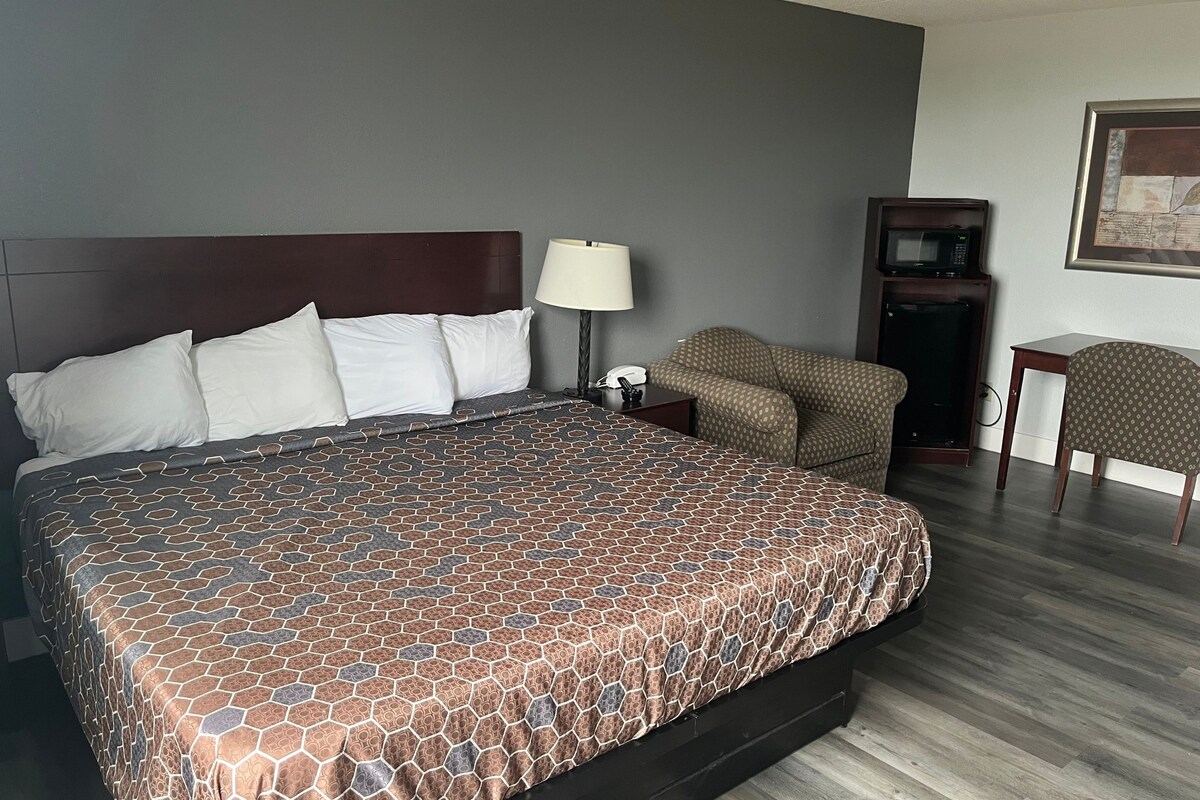 A hotel room with a large bed, a bedside table with a lamp, a chair, a desk with a computer, and a microwave.