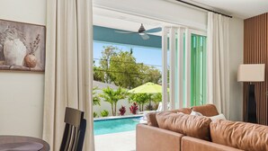 Un salon avec un canapé, un tableau, un ventilateur de plafond et une vue sur une piscine et la verdure environnante.