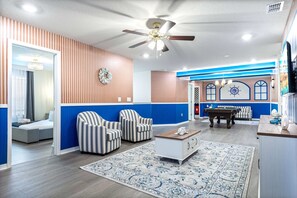 A living room with striped armchairs, a coffee table, a blue accent wall, a ceiling fan, and a billiards table.