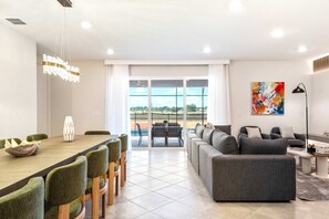Un salon moderne avec un coin repas, un canapé gris et une vue sur un court de tennis grâce à de grandes fenêtres.