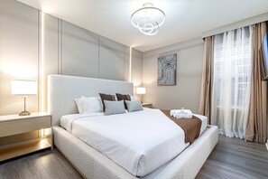 A modern hotel room with a large bed, a bedside table with a lamp, a wall-mounted artwork, and a view of the city through sheer curtains.