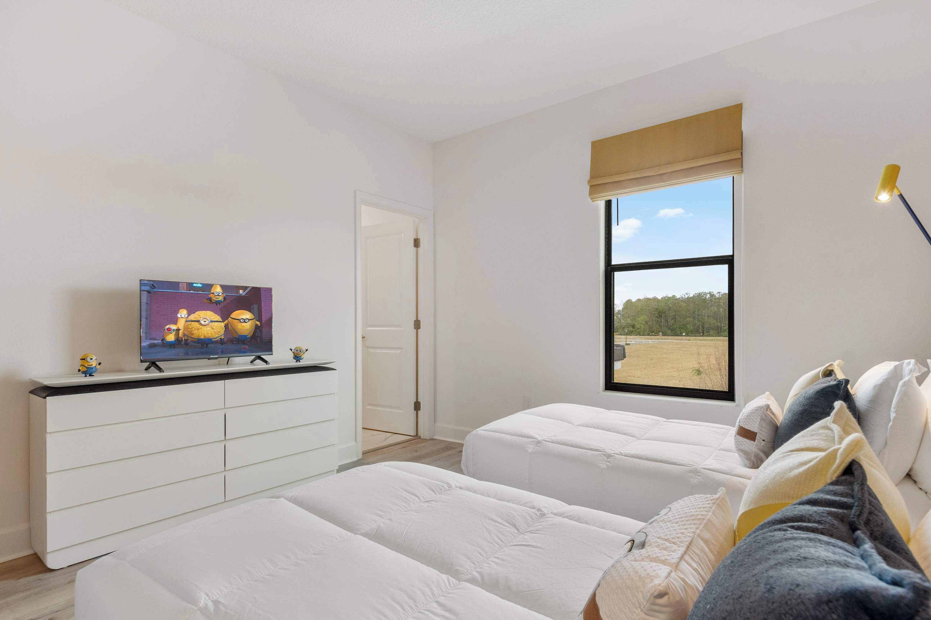 A modern bedroom with two beds, a TV on a white dresser, a large window with a view of a field, and a door leading to another room.