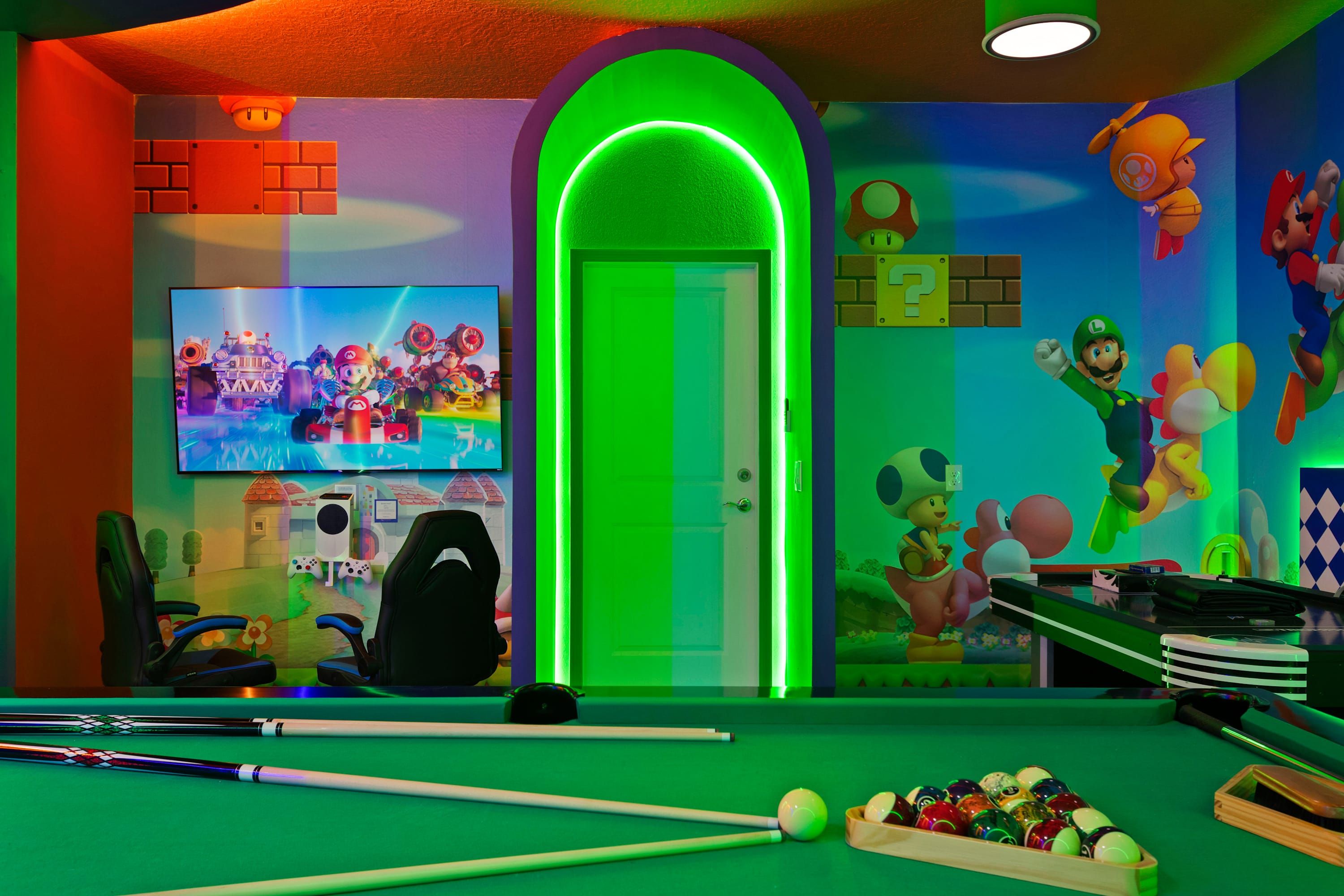A gaming room with a pool table, gaming chairs, and a TV displaying a Mario-themed game.