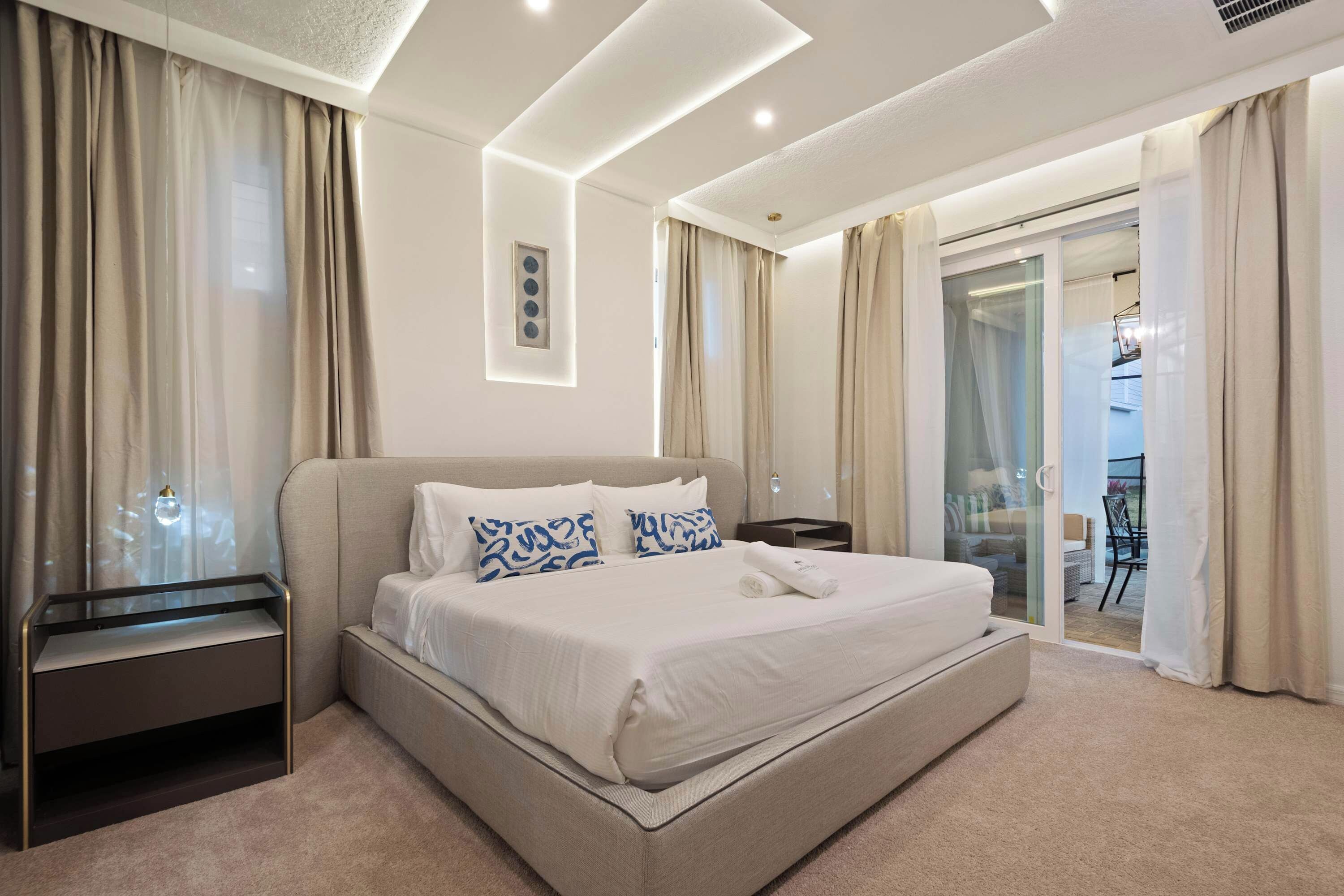 A modern hotel room with a large bed, a bedside table, a glass desk, and a sliding glass door to the balcony.