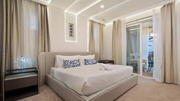 A modern hotel room with a large bed, a bedside table, a glass desk, and a sliding glass door to the balcony.