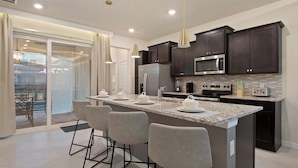 A modern kitchen with dark cabinets, a large island with bar stools, and a sliding glass door leading to a patio. - Luxurious Windsor Cay Villa Private Pool, Epic Game Room & Minutes to Disney! (Clermont)