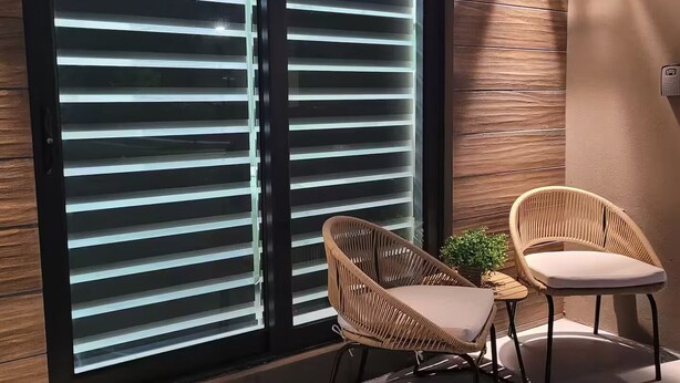 Un espace de détente extérieur moderne, avec deux fauteuils en osier et une petite table, situé à côté d’une grande fenêtre dotée de stores horizontaux.