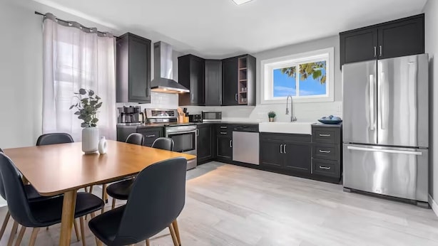 A modern kitchen with dark cabinets, a stainless steel refrigerator, and a dining area with a wooden table and chairs. - Spacious 3BDs - Close to Wineries, Brock & Falls! (Thorold)
