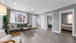 A spacious living room with a green sofa, a wooden coffee table, and a beige armchair. There is a large window with curtains, a door leading to another room, and a view of a house outside. - Spacious 3BDs - Close to Wineries, Brock & Falls! (Thorold)