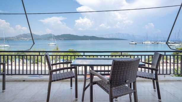 Una terraza con mobiliario de mimbre y una mesa, con vistas a un puerto deportivo con barcos y montañas a lo lejos.