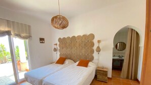Un lit double avec deux oreillers, une tête de lit en osier, une table de chevet avec une lampe, et une vue sur la salle de bain à travers une arche.