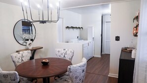 Uma sala de jantar com uma mesa redonda e cadeiras estampadas, um lustre, um espelho e uma kitchenette ao fundo.