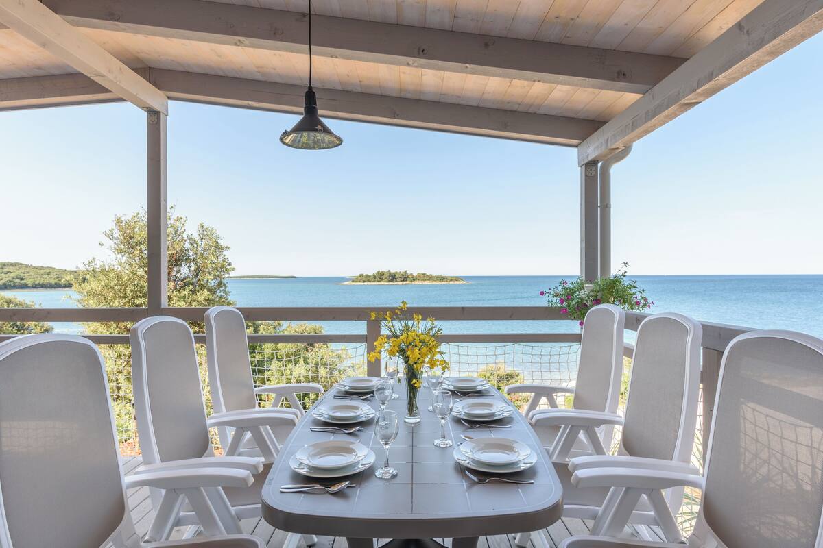 Een overdekte eethoek met een tafel voor zes personen, met uitzicht op zee en een eiland in de verte.