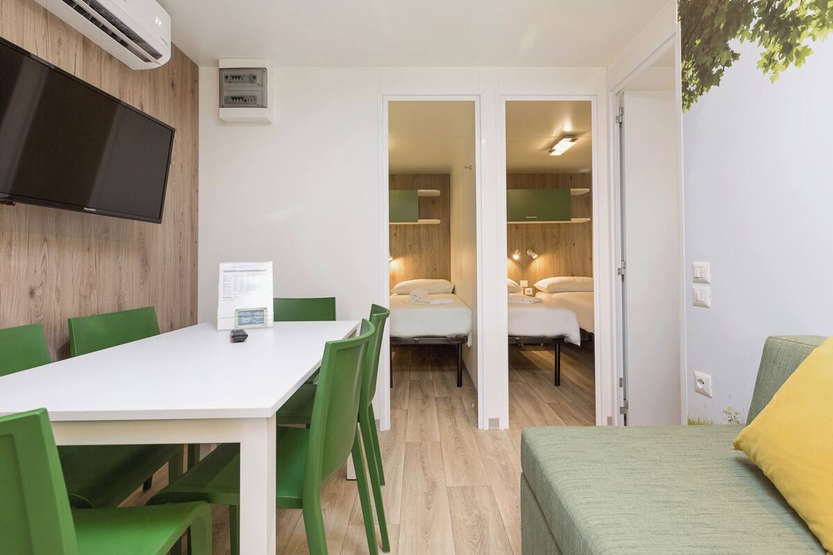 Een compacte kamer met een witte tafel, groene stoelen, een bed met een groen en geel kussen en een televisie die aan de muur is bevestigd.