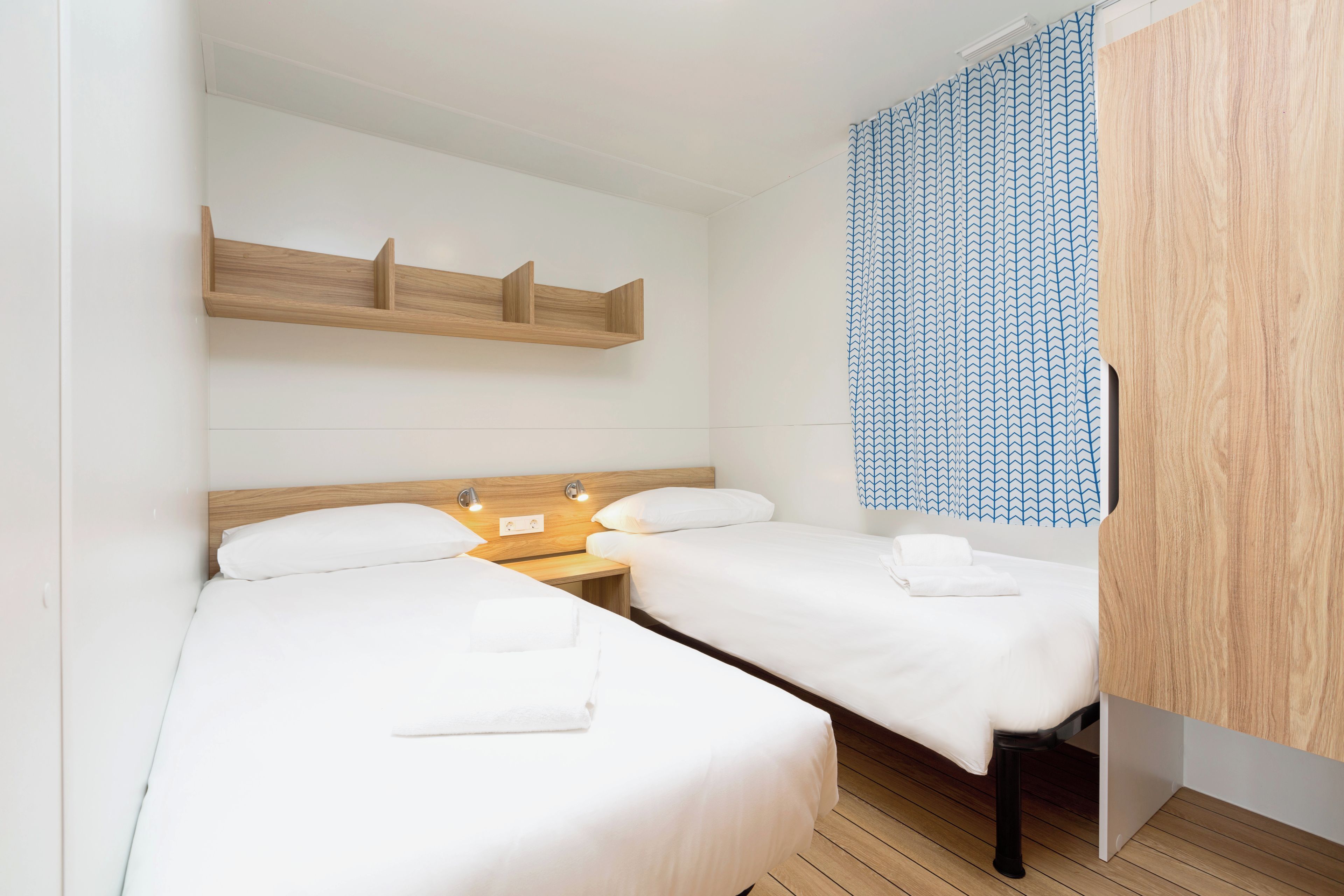 A hotel room with two single beds, a wooden headboard, a shelf above the beds, and a patterned curtain.