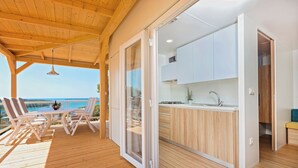 Eine Holzterrasse mit Tisch und Stühlen, eine Küche mit weißen Schränken und Blick aufs Meer.
