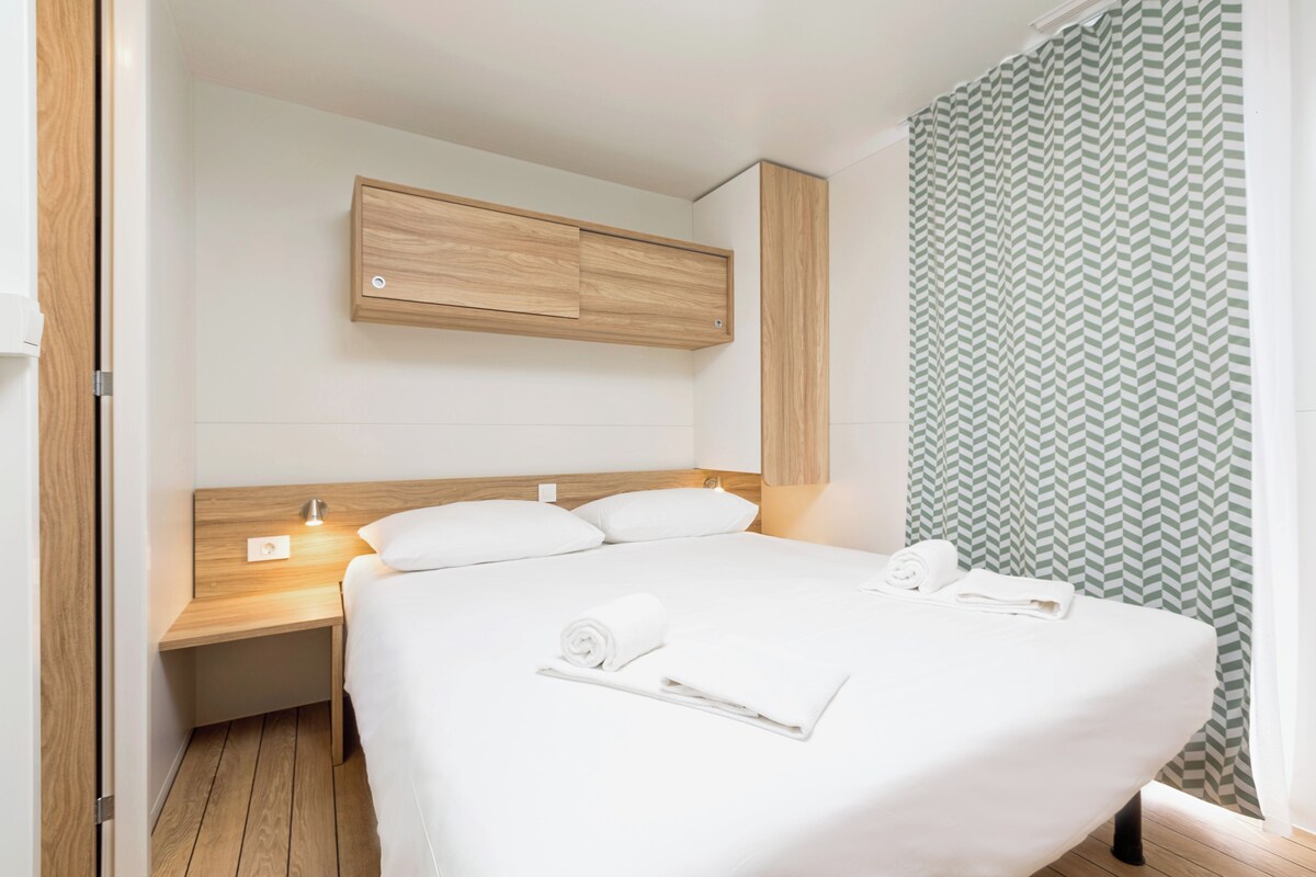 Une chambre d’hôtel moderne avec une tête de lit en bois, une table de chevet avec une lampe, un grand lit avec du linge de lit blanc et un rideau de douche à motifs verts.