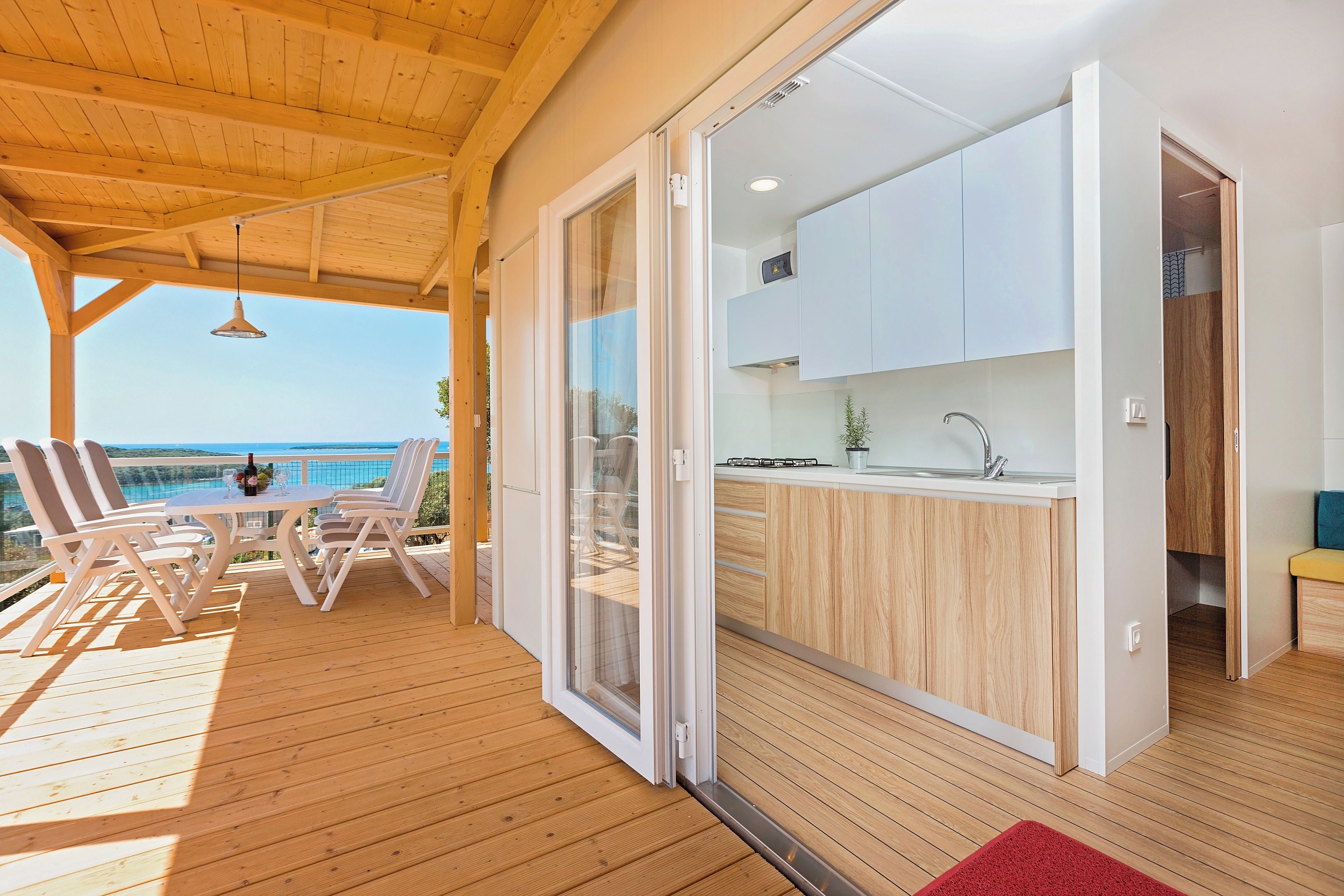 Een houten terras met een tafel en stoelen, een keuken met witte kasten, en uitzicht op zee.