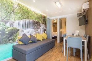 Une pièce avec un canapé gris, des coussins jaunes et gris, une table à manger avec des chaises, et un téléviseur fixé au mur.