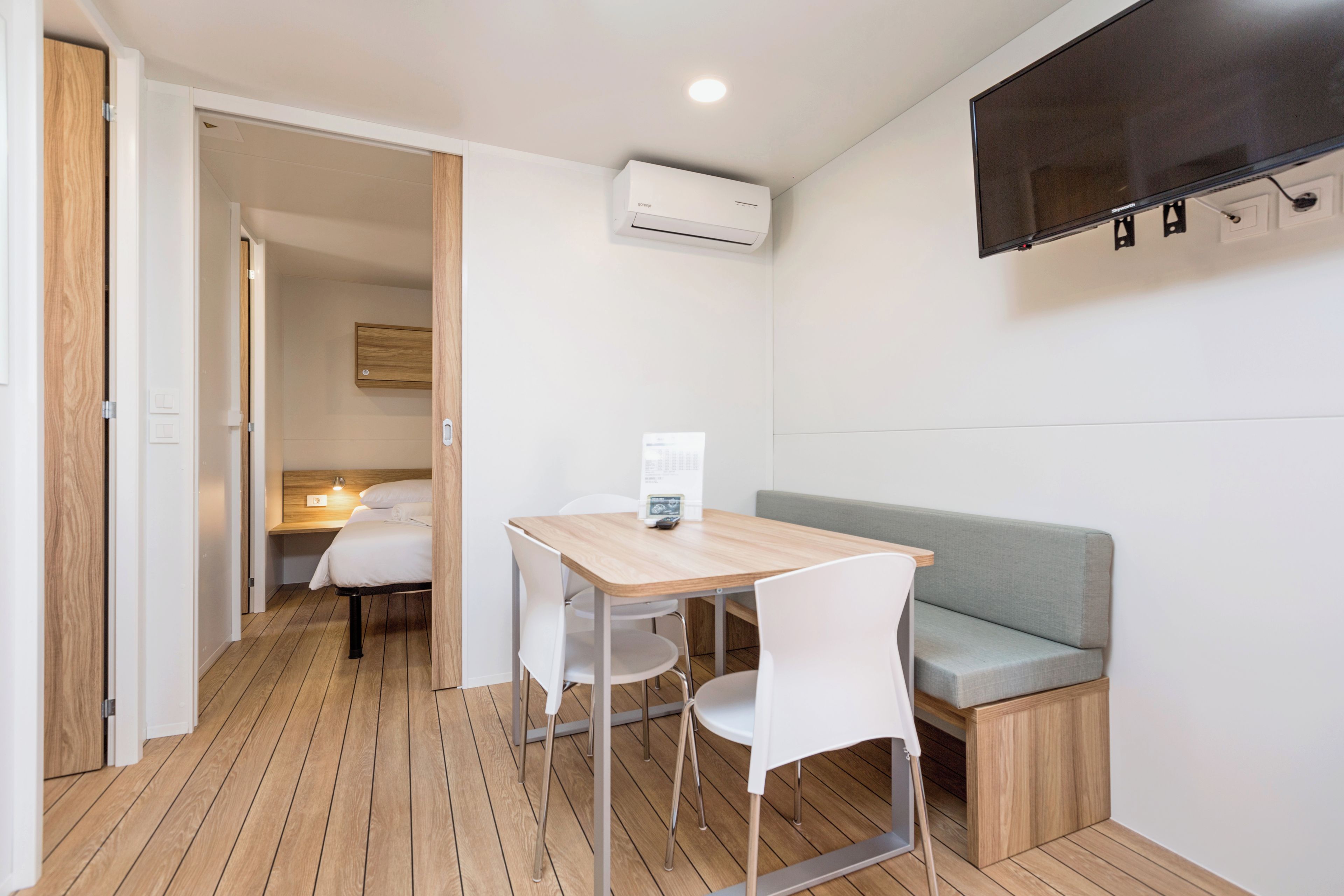 A compact room with a wooden floor, a small table with chairs, a sofa, a flat-screen TV mounted on the wall, and an air conditioning unit.