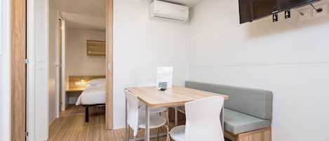 A compact room with a wooden floor, a small table with chairs, a sofa, a flat-screen TV mounted on the wall, and an air conditioning unit.