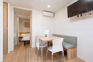 Une pièce de taille modeste avec un plancher en bois, une petite table avec des chaises, un canapé, un téléviseur à écran plat fixé au mur et un appareil de climatisation.