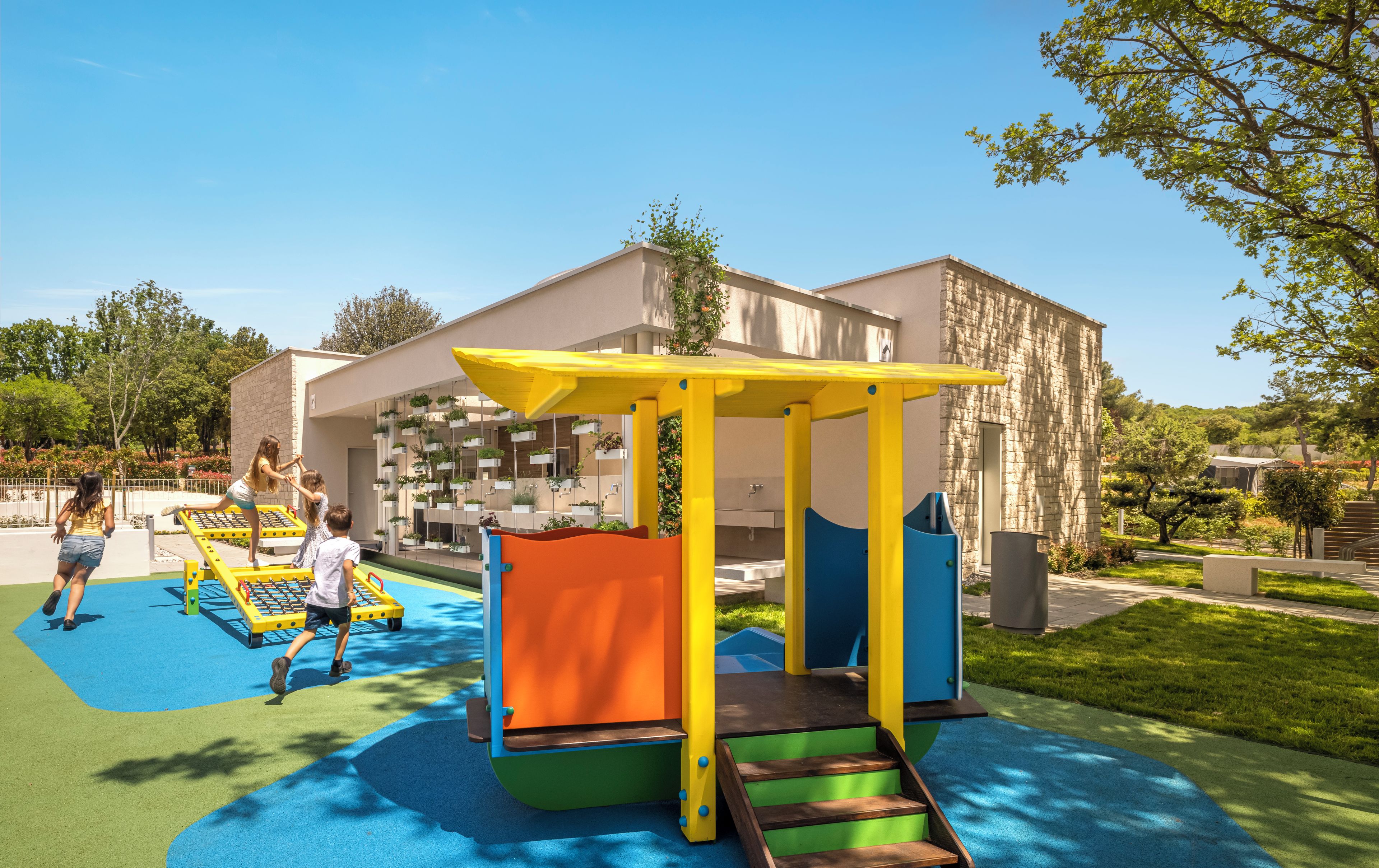 Un terrain de jeux avec des structures colorées, un bâtiment à la façade en pierre et une pelouse bien entretenue.