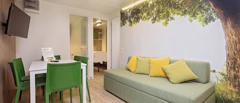 Una habitación con un sofá verde, una mesa de comedor con sillas, un televisor de pantalla plana montado en la pared y un mural de un gran árbol en la pared.