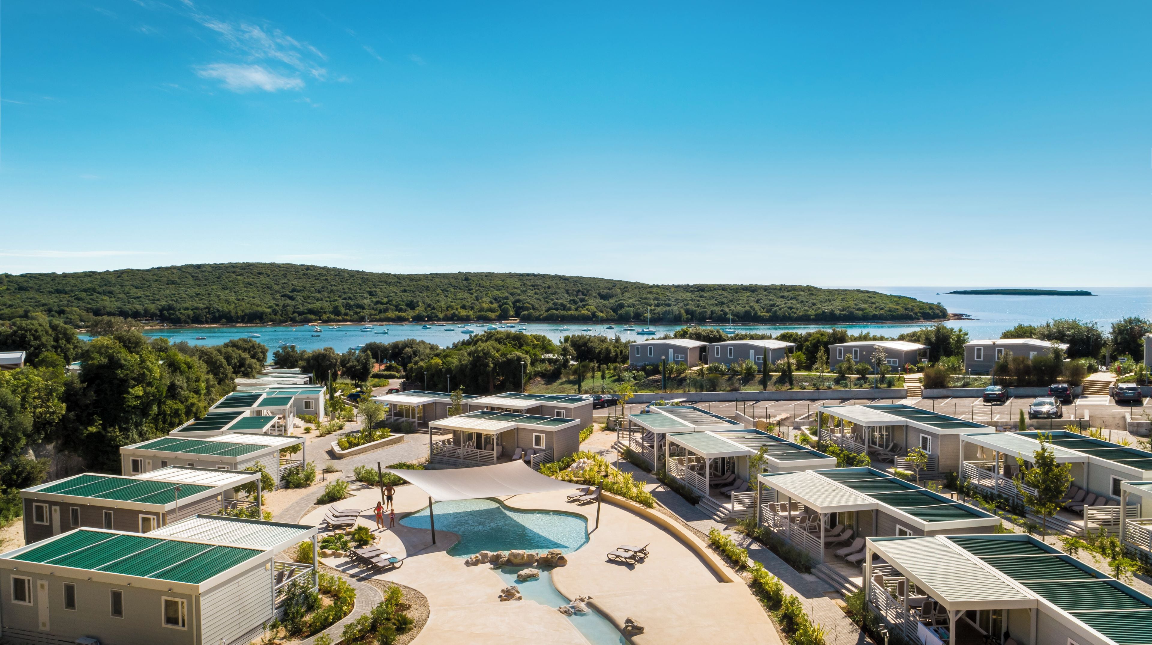 Une station balnéaire proposant plusieurs types d’hébergements en style bungalow, une piscine centrale et une vue sur la mer et les collines.