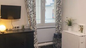 Une petite chambre comprenant un lit, un bureau, une télévision et une fenêtre agrémentée de rideaux à motifs.