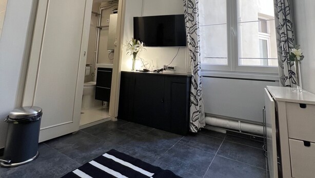 Un espace de vie compact comprenant un coin cuisine, un tapis noir et blanc, un petit bureau et une fenêtre agrémentée de rideaux à motifs.