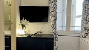 Une chambre à coucher avec un lit, un téléviseur fixé au mur, une commode, une fenêtre avec des rideaux et une salle de bain visible à travers une porte ouverte.