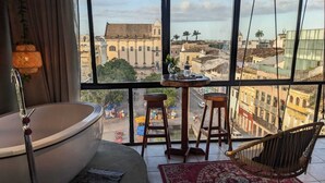 Una habitación con una bañera grande, una mesa redonda con taburetes, una silla y vistas a un paisaje urbano a través de grandes ventanales.