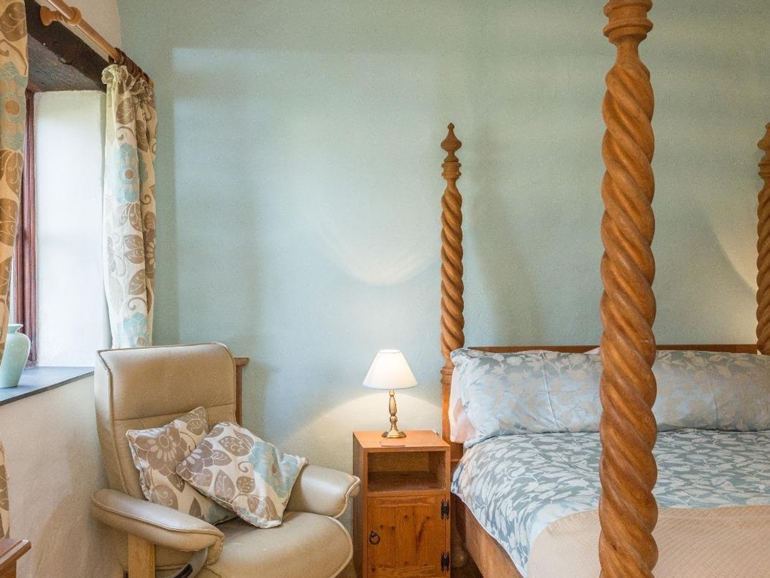 Une chambre à coucher avec un lit à baldaquin, un fauteuil avec un coussin, une table de chevet avec une lampe et une fenêtre avec des rideaux.