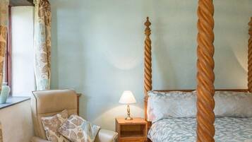 Une chambre à coucher avec un lit à baldaquin, un fauteuil avec un coussin, une table de chevet avec une lampe et une fenêtre avec des rideaux.
