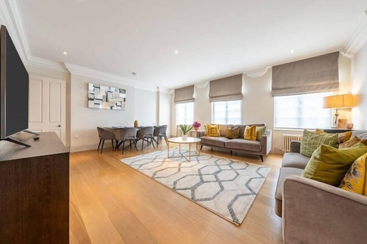 Un salon moderne avec un canapé, une table basse et des fauteuils. On y trouve un téléviseur posé sur un meuble en bois, un élément décoratif mural et une fenêtre avec des rideaux.