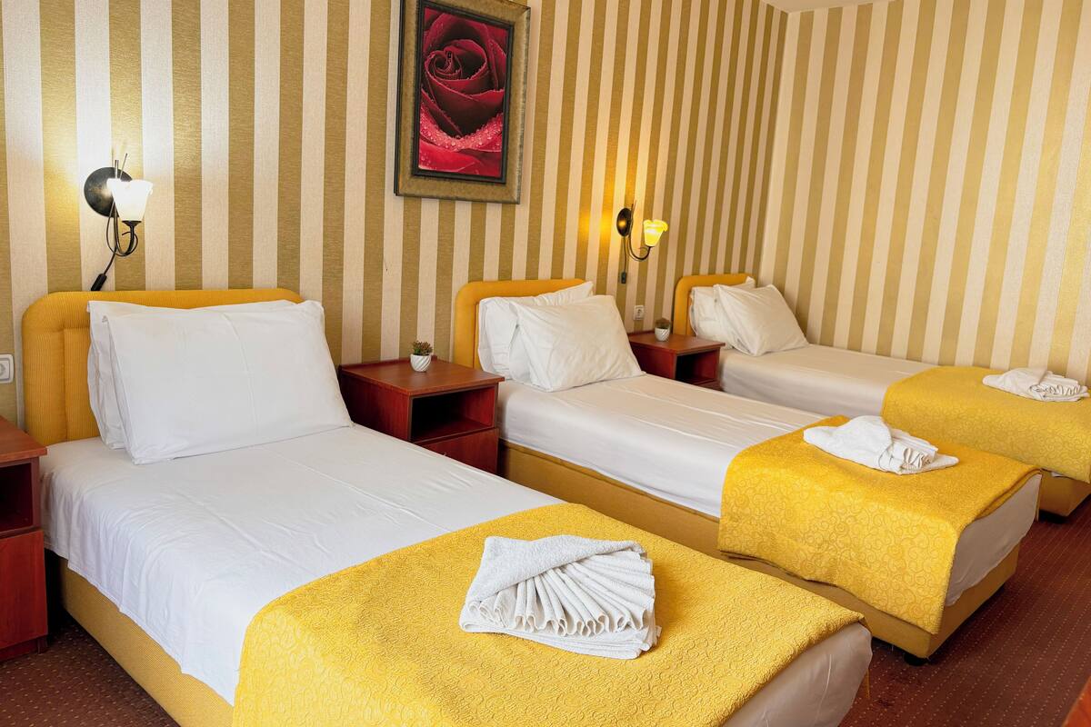 Une chambre d’hôtel avec trois lits, chacun recouvert d’une couvre-lit jaune et doté de draps blancs. Il y a deux tables de chevet avec des lampes, un tableau encadré au mur et une petite plante sur l’une des tables.