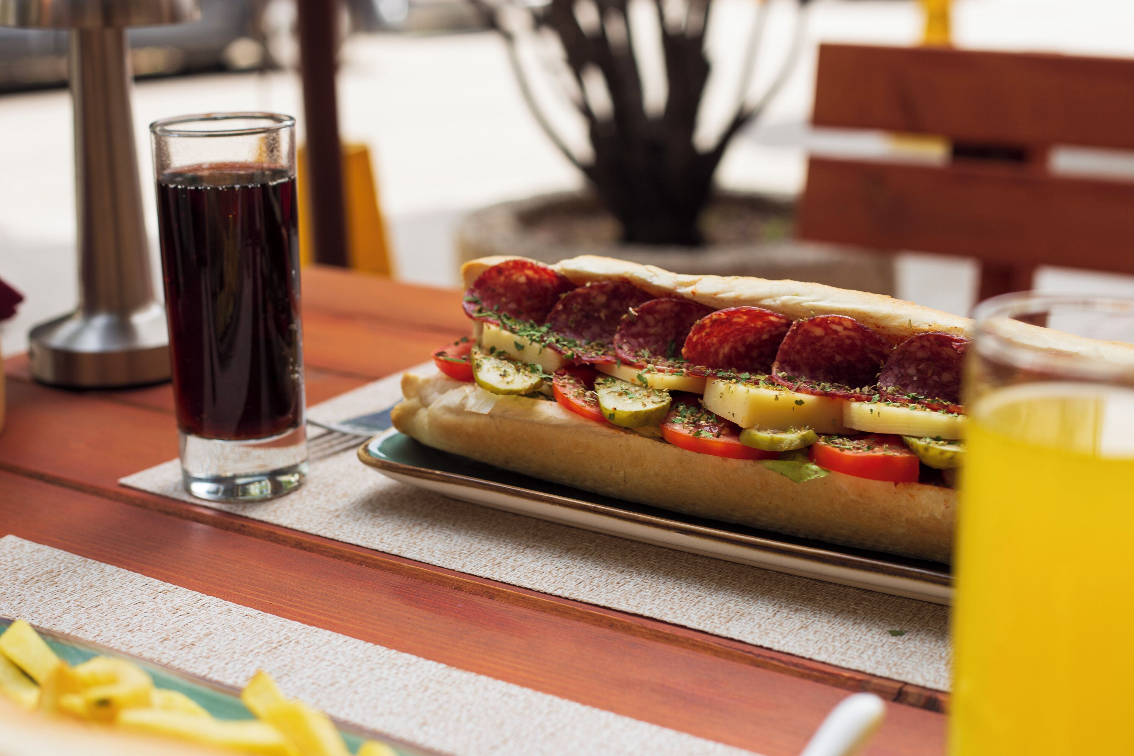 A sandwich with various toppings on a plate, a glass of dark liquid, and a glass of yellow liquid on a wooden table.