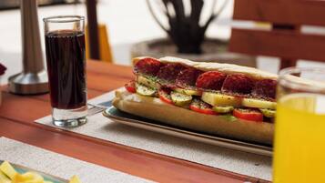 A sandwich with various toppings on a plate, a glass of dark liquid, and a glass of yellow liquid on a wooden table.