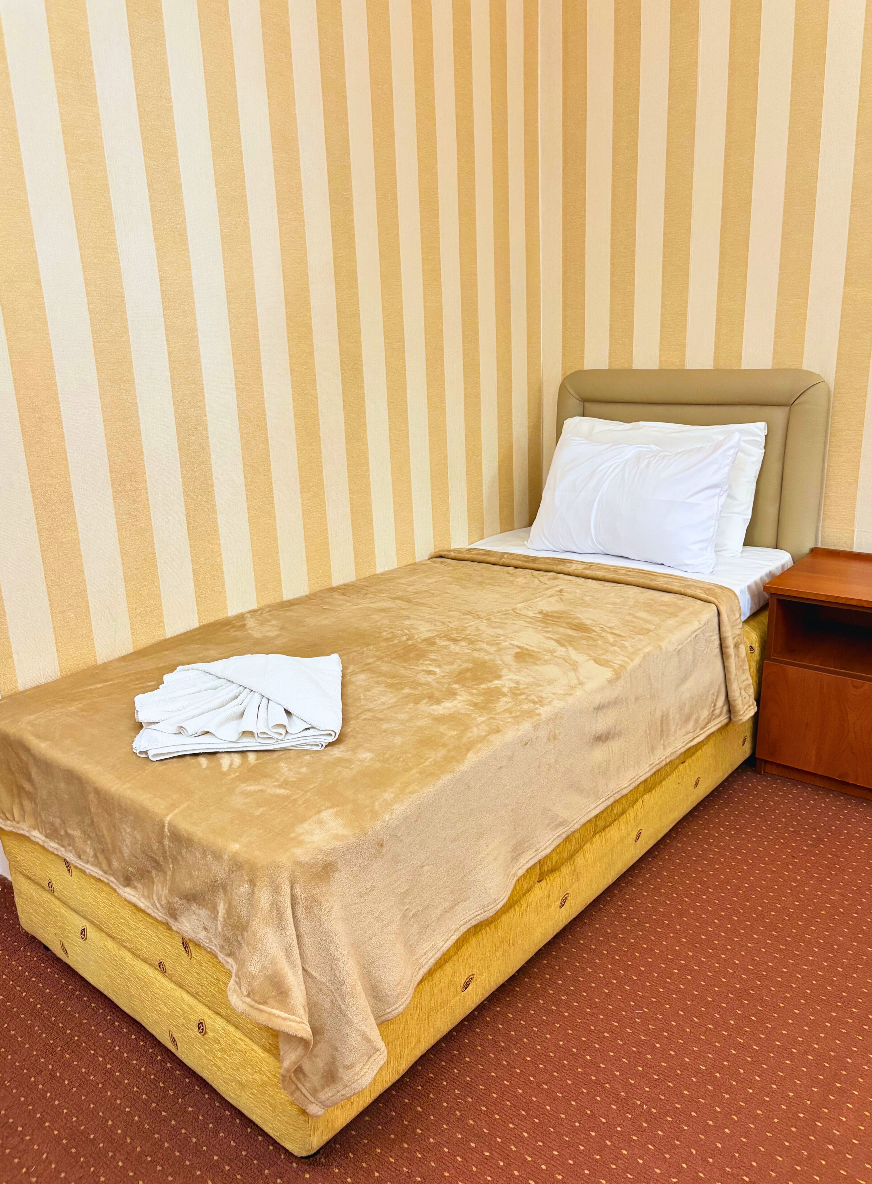 Een eenpersoonsbed met een beige deken, witte kussens en een opgevouwen witte handdoek op het voeteneinde. De kamer heeft een gestreept behang en een houten nachtkastje.